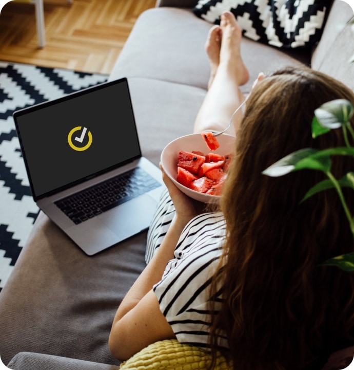 Женщина на диване ест фрукты, рядом с ней лежит ноутбук с логотипом Norton